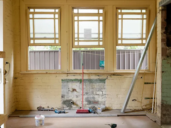 Old kitchen under renovation with exposed pipes, tools, and a broom against a brick wall